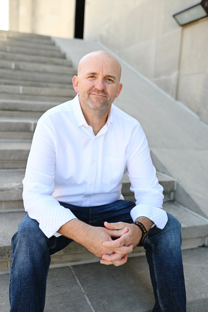 Man sitting on steps wearing a white shirt and blue jeans, with a neutral background